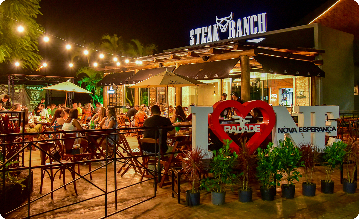 Fachada iluminada da churrascaria Steak Ranch à noite com clientes no espaço externo